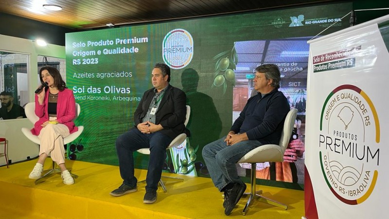 Num palco de piso amarelo e de fundo composto por um painel verde, Simone está sentada ao lado de dois homens, também sentados em cadeiras brancas. Ela fala ao microfone e traja um blazer rosa. O painel verde ao fundo conta com os escritos Selo Produto Premium e Qualidade RS 2023. À direita na imagem, há um pôster com o símbolo do selo.