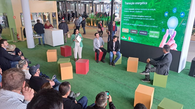 Pessoas em uma arquibancada assistem ao seminário. Na frente um telão verde com informações sobre transição energética. Entre o telão e a arquibancada estão os palestrantes: três homens sentados em caixas coloridas e uma mulher está em pé, falando.