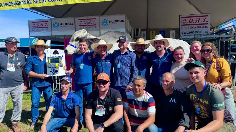 Na imagem, um grupo de aproximadamente 15 pessoas posa para foto em frente a um estande na Expointer. Entre os participantes, est&atilde;o homens e mulheres, alguns usando chap&eacute;us de palha ou bon&eacute;s, e muitos com crach&aacute;s de identifica&ccedil;&atilde;o pendurados no pesco&ccedil;o. Ao fundo, &eacute; poss&iacute;vel ver um grande boi branco da ra&ccedil;a zebu&iacute;na, identificado por uma faixa azul na cabe&ccedil;a, em destaque como atra&ccedil;&atilde;o principal. Um dos homens segura um painel eletr&ocirc;nico que marca &ldquo;1430&rdquo;, indicando o peso do animal, que simboliza o recorde de maior bovino j&aacute; apresentado na feira. O ambiente &eacute; de celebra&ccedil;&atilde;o e registro hist&oacute;rico.