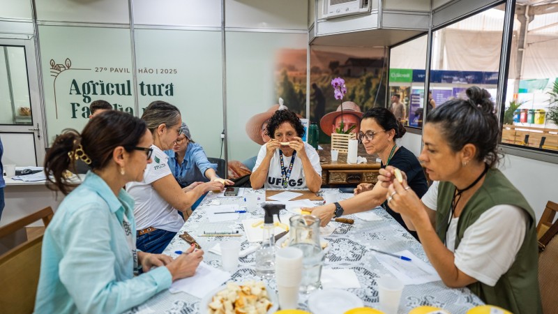 Na imagem, um grupo de seis mulheres est&aacute; reunido em uma mesa retangular, participando de uma atividade de degusta&ccedil;&atilde;o de queijos. Elas seguram peda&ccedil;os do produto, avaliando aroma, sabor e textura, enquanto fazem anota&ccedil;&otilde;es em folhas de papel. A mesa est&aacute; coberta com toalha branca rendada e sobre ela h&aacute; queijos inteiros numerados, peda&ccedil;os cortados em pratos e copos pl&aacute;sticos. Ao fundo, o espa&ccedil;o &eacute; identificado como parte do 27&ordm; Pavilh&atilde;o da Agricultura Familiar da Expointer 2025, com pain&eacute;is ilustrativos e logotipo vis&iacute;vel.