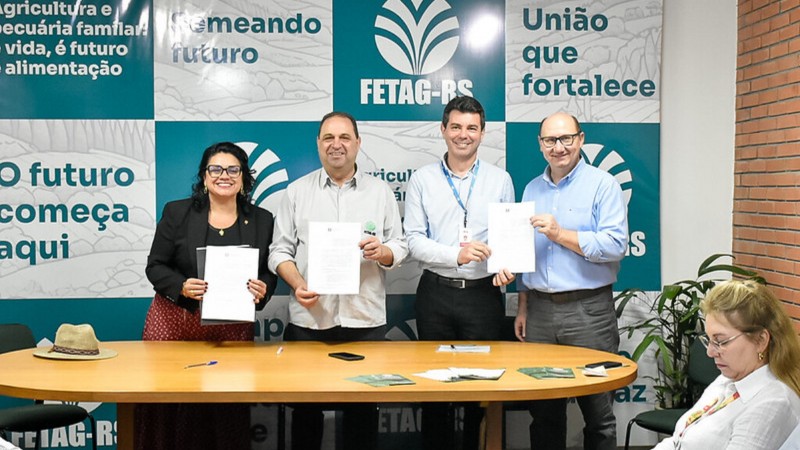 A imagem mostra quatro pessoas em p&eacute; atr&aacute;s de uma mesa de madeira, segurando documentos assinados e sorrindo para a c&acirc;mera. Eles est&atilde;o participando de um evento formal relacionado &agrave; agricultura familiar. Ao fundo, h&aacute; um painel com logotipos e frases institucionais, como "FETAG-RS", "Uni&atilde;o que fortalece", "Agricultura e pecu&aacute;ria familiar &eacute; vida, &eacute; futuro, &eacute; alimenta&ccedil;&atilde;o" e "O futuro come&ccedil;a aqui".

As quatro pessoas no centro da imagem s&atilde;o tr&ecirc;s homens e uma mulher. A mulher est&aacute; &agrave; esquerda, vestindo um blazer preto, &oacute;culos e saia vermelha com estampa. &Agrave; direita dela, os tr&ecirc;s homens est&atilde;o vestidos com camisas sociais, um deles com crach&aacute;. Todos seguram folhas de papel, aparentemente contratos ou acordos assinados.

&Agrave; direita da imagem, uma mulher loira est&aacute; sentada e concentrada em um celular. H&aacute; uma planta decorativa ao fundo e parte de uma parede de tijolos aparentes &agrave; direita.