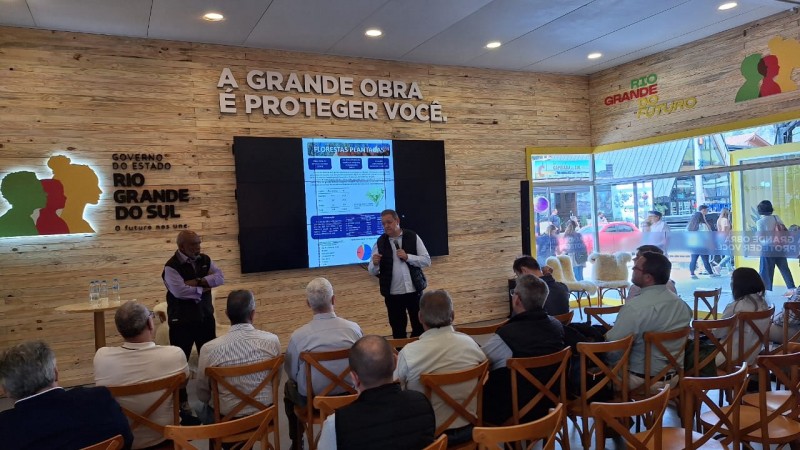 Foto de uma sala dentro do Parque Assis Brasil, tirada do fundo. Um homem aparece de p&eacute;, falando ao microfone, com v&aacute;rias pessoas assistindo sentadas. H&aacute; uma tela com apresenta&ccedil;&atilde;o atr&aacute;s dele, e uma logo do governo na parede.