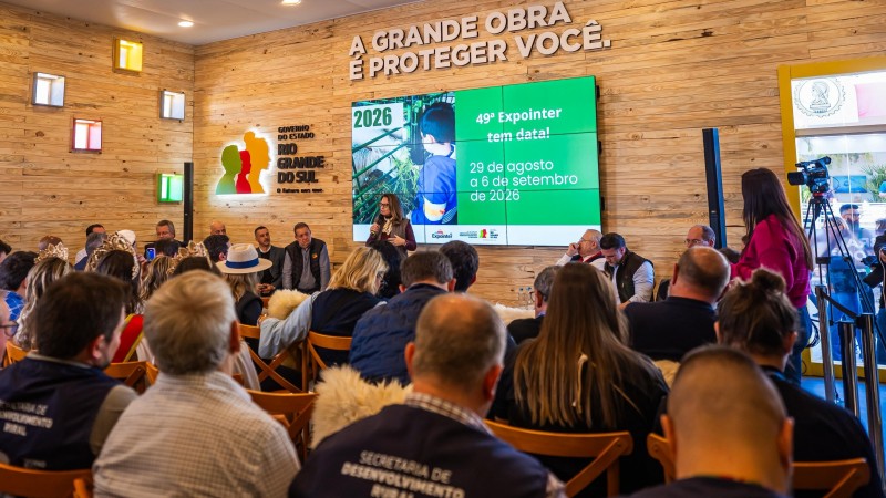 Foto mostra a coletiva da Expointer, com p&uacute;blico sentado acompanhando apresenta&ccedil;&atilde;o. No tel&atilde;o, as datas de 2026. A parede do local &eacute; em madeira clara. Acima do tel&atilde;o a frase "A grande obra &eacute; proteger voc&ecirc;" e a logomarca do RS.