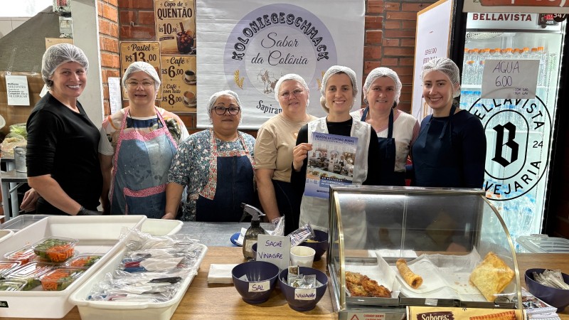 Na 49&ordf; Expointer, sete integrantes da Koloniegeschmack apresentam produtos e receitas da gastronomia colonial aos visitantes da feira. Elas est&atilde;o em frente a uma mesa com produtos coloniais e segurando uma revista