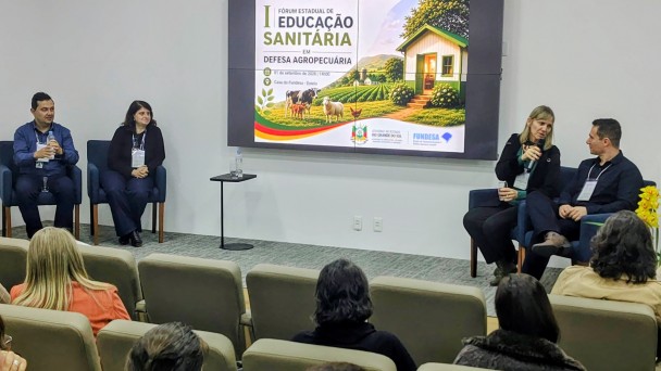 Foto mostra painelistas sentados em poltronas no palco, em frente a plateia. Ao fundo, uma tela com a apresenta&ccedil;&atilde;o do tema sobre educa&ccedil;&atilde;o sanit&aacute;ria. 