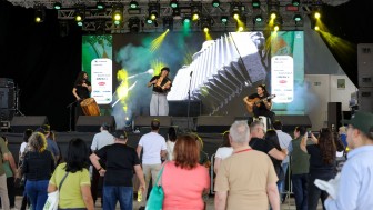 Fotografia de um show musical em um palco iluminado por luzes amarelas e verdes. Ao centro, um m&uacute;sico toca acorde&atilde;o; &agrave; esquerda, outro toca um tambor, e &agrave; direita, um terceiro m&uacute;sico toca viol&atilde;o sentado em um banco. Ao fundo, um grande painel de LED exibe uma imagem ampliada de um acorde&atilde;o. Na frente do palco, h&aacute; uma plateia numerosa, de costas para a c&acirc;mera, acompanhando a apresenta&ccedil;&atilde;o. A estrutura met&aacute;lica do palco est&aacute; repleta de refletores e equipamentos de ilumina&ccedil;&atilde;o, e h&aacute; fuma&ccedil;a c&ecirc;nica espalhada pelo ambiente.