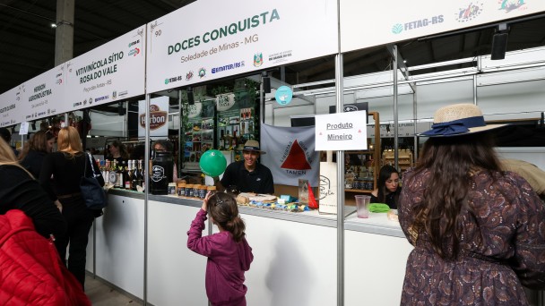 Foto mostra bancas num corredor do Pavilh&atilde;o da Agricultura Familiar. Numa delas, ao fundo, se v&ecirc; a bandeira do Estado de Minas Gerais, com o fundo branco e um tri&acirc;ngulo vermelho no centro. H&aacute; uma menina na frente da banca com um bal&atilde;o verde. Outras mulheres est&atilde;o visitando outras bancas. 