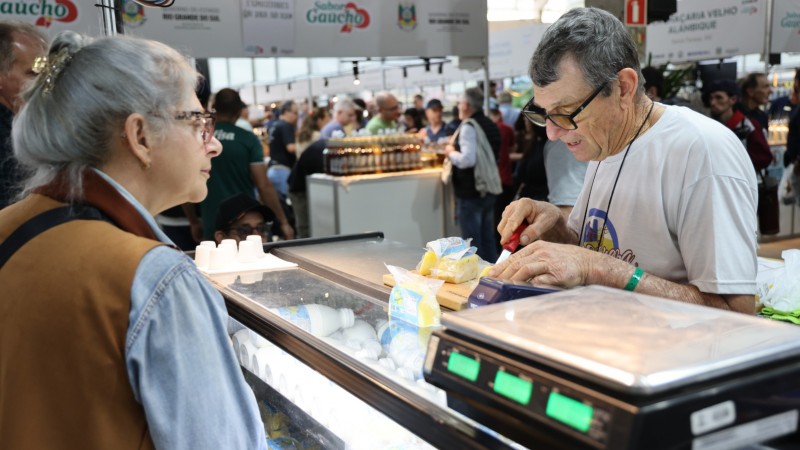 Foto mostra um expositor cortando um queijo atr&aacute;s do balc&atilde;o. Uma mulher de &oacute;culos e cabelos brancos est&aacute; parada observando atentamente a explica&ccedil;&atilde;o do expositor. Ao lado dele, h&aacute; uma balan&ccedil;a digital. Ao fundo, aparece o corredor com v&aacute;rias bancas lotadas de p&uacute;blico circulando pelos corredores do pavilh&atilde;o.