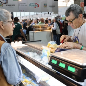 Foto mostra um expositor cortando um queijo atr&aacute;s do balc&atilde;o. Uma mulher de &oacute;culos e cabelos brancos est&aacute; parada observando atentamente a explica&ccedil;&atilde;o do expositor. Ao lado dele, h&aacute; uma balan&ccedil;a digital. Ao fundo, aparece o corredor com v&aacute;rias bancas lotadas de p&uacute;blico circulando pelos corredores do pavilh&atilde;o.